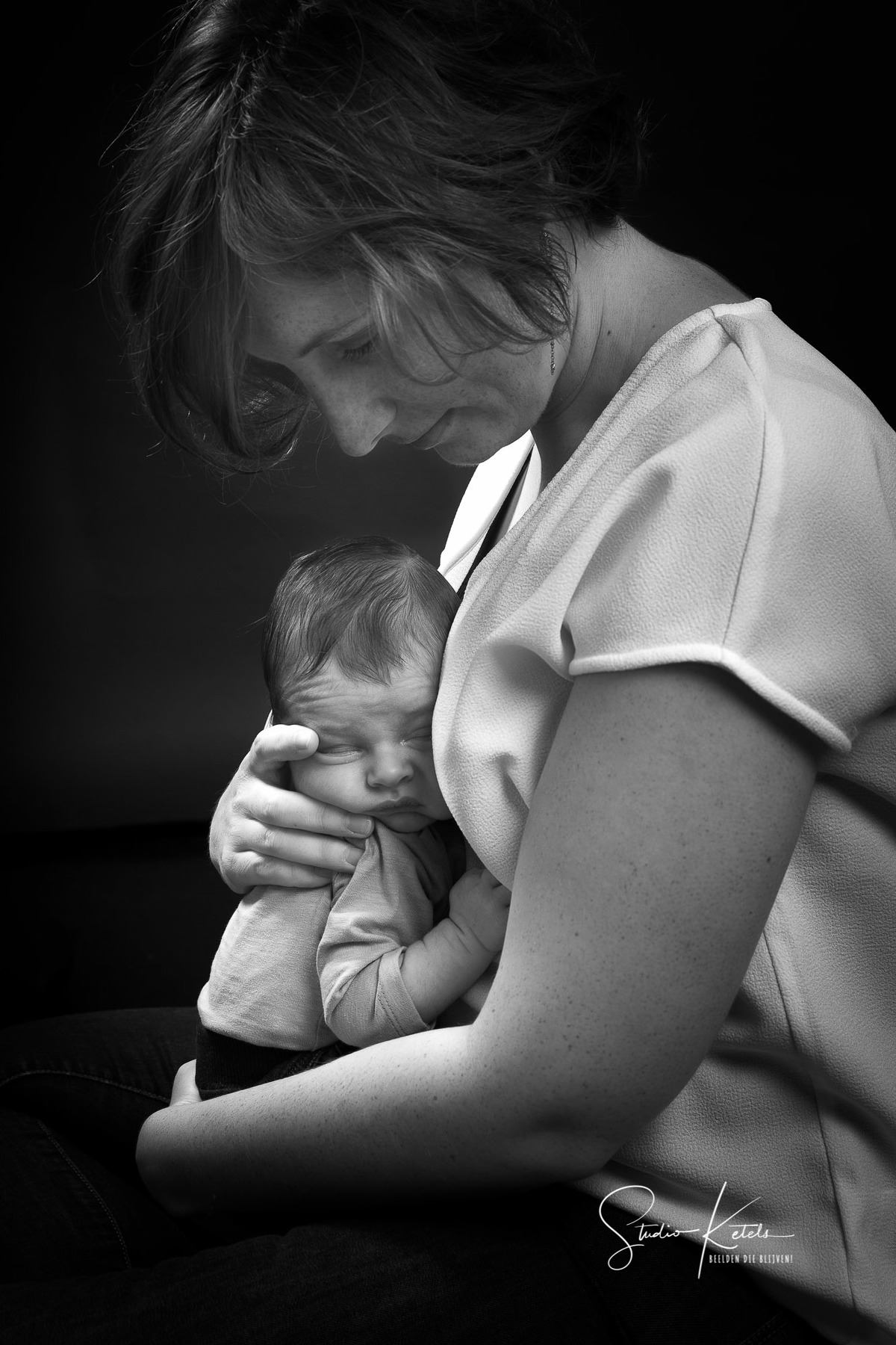 Intiem zwart-wit-portret van een pasgeboren babytje in de armen van de moeder. Zij kijkt beschermend naar haar babytje, dat ze tegen zich aan houdt. Portret door Studio Ketels